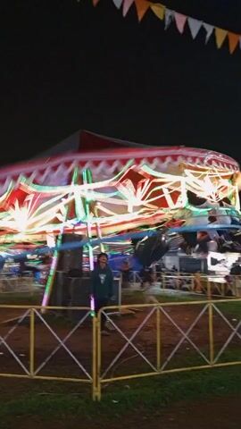 At night, a colorfully lit carousel spins rapidly in an open-air setting, possibly a carnival or fair. The carousel has a red and white striped roof and multiple levels of lights. A yellow metal fence surrounds the ride. A person stands outside the fence. Above, a string of pennants is strung across the scene. Other fairground attractions can be seen in the background.