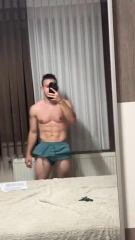 A male, shirtless with green shorts, stands in front of a mirror, filming himself with a phone. He flexes his bicep and displays his abs. The room has brown and white curtains, and a bed with a white comforter is visible in the foreground.