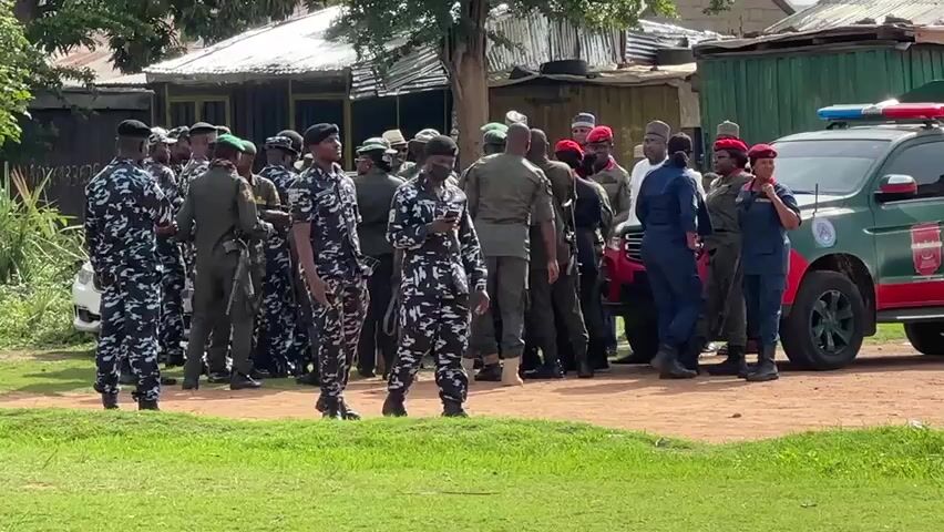 The video shows a gathering of uniformed individuals. Some are in blue and gray camouflage, others in olive green, and some in blue uniforms. A few individuals wear red berets. A green SUV with red and blue lights on top is parked nearby. The people appear to be in a yard with green grass. Buildings can be seen in the background. Some individuals are talking to each other, while one person is looking at their phone.
