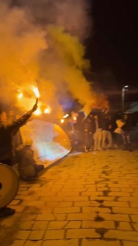 At night, a group of people celebrates in a narrow brick-paved street. Some hold burning flares, creating orange and white smoke. Two people sit on the ground holding a banner with illegible words. A drum sits next to them. The crowd is mostly men, dressed in casual dark clothing. A brick wall lines one side of the street.