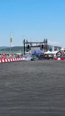 A white BMW drifting in a circle, emitting smoke. A crowd of spectators is visible behind a red and white barrier. A stage with speakers and tents is set up in the background. The sky is clear and blue.