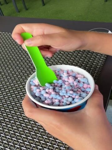 A person's hands are holding a white cup filled with small, bite-sized pieces of pink and blue frozen dessert. The dessert pieces are shaped like tiny flowers or clovers. A bright green spoon is being used to scoop the dessert from the cup. The person is lifting a spoonful of the dessert towards their mouth. The cup has a red and blue design on its exterior. The dessert is sitting on a dark, woven placemat with a diamond pattern. In the blurred background, there are hints of outdoor seating with green elements, suggesting an outdoor dining area. The lighting indicates it is daytime. The scene appears casual and suggests someone is enjoying a treat. No visible text is present on the cup or the surrounding objects. The location context is La Vila Joiosa, Spain.