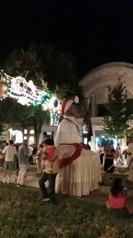 A nighttime scene shows a large, doll-like figure dressed in a white gown with red trim and a red and white headpiece. The figure is seated and appears to be a puppet. A crowd of people is gathered around, some watching the figure and others looking at decorations strung across the street. The decorations include lights and circular frames containing images, one of which shows a child. The area appears to be an outdoor public space, possibly a street or plaza, with trees and buildings in the background. The overall atmosphere seems festive.
