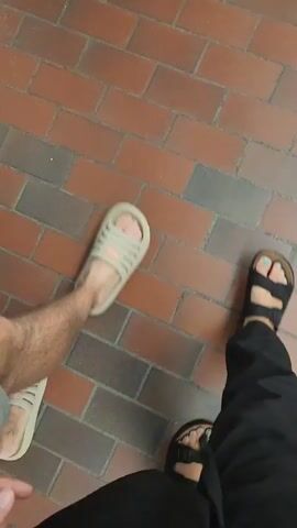 Two people stand on a brick floor.  One person wears beige slide sandals on both feet. The other person wears black sandals with a blue pedicure. The camera appears to be held at chest level by the person wearing the black sandals. A gray wall is on one side.