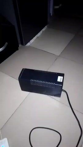 The video shows a black uninterruptible power supply (UPS) on a tiled floor. The UPS has a power cord connected to it. Initially, an electronic buzzing sound is audible. A hand briefly enters the frame. The camera focuses on the top and side of the UPS, displaying its ventilation slats and a sticker. The buzzing sound persists.