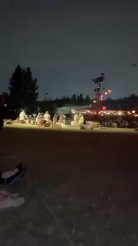 The video shows an outdoor event at night, likely a performance or gathering, observed from the perspective of someone sitting on the ground. In the distance, figures in what appear to be white robes sit or kneel on a brightly lit grassy area. Behind them are more people, lit by strings of lights and a tower with stage lights. The sky is dark and cloudy. In the foreground, the legs and feet of the person recording the video are visible, clad in shoes of different colors.