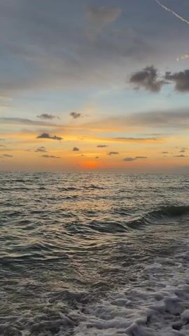 The video shows a sunset over the ocean. The sun is setting on the horizon, casting a warm orange glow across the sky. The clouds are illuminated by the setting sun, with streaks of yellow and pink visible. The ocean is dark and choppy, with waves crashing on the shore in the foreground. The scene is peaceful and serene.