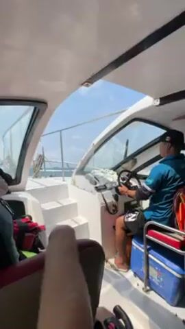 The interior of a small motorboat with a white fiberglass hull is shown. A man in a turquoise shirt and black shorts, wearing a black baseball cap, is steering the boat.  A red and white cooler sits next to him. Other people are inside the boat, and there is a glimpse of legs in the foreground.  The sky is blue and bright, and the ocean is visible through the windshield.