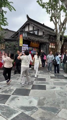 The video captures a busy outdoor scene in what appears to be a traditional Chinese street. People of varying ages walk and interact, many holding bags or phones. There's a food stall with a menu display visible, and several people are gathered around it. The buildings have traditional Chinese architectural features, like dark roofs and intricate wooden detailing. The ground is paved with a checkered pattern of black and gray stones. The overall atmosphere is lively, with sounds of chatter and activity in the background. The lighting indicates it's daytime, possibly overcast.
