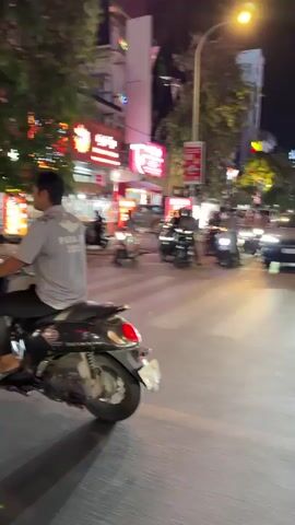 A street scene at night. Many motorcycles are stopped at a crosswalk. Several buildings with bright lights and signs are visible in the background. A traffic light is also visible. The camera is moving.