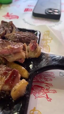The video shows a plate of sliced steak and boiled potatoes on a table. The steak is rare to medium-rare, with some pieces showing a pink interior. The potatoes are small and yellow, glistening with moisture. The plate is black and sits on a tablecloth with a red and white pattern. A smartphone and a green bottle are visible in the background. The camera focuses on the food, panning slightly to show the arrangement of the steak and potatoes. The voice is likely that of a speaker, describing the portion of the steak and potato as "small", when compared to another time that a larger portion was served.