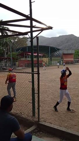 The video shows a baseball field on a cloudy day with mountains in the background. Several people, presumably baseball players, are scattered around the field. Some are running the bases, while others are standing and watching. One player is seen giving a high-five to another. A man in a blue and green jersey with the name "J. Bermudez" and the number "39" is also visible. The field is surrounded by a chain-link fence, and there is a covered seating area visible in the background. A building is visible to the right. The general atmosphere suggests a casual baseball game or practice session.