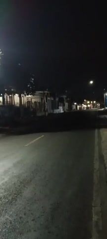 The video pans from left to right across a dimly lit street at night. There are houses and buildings lining the street, with some lights visible from windows and street lamps. A motorcycle is parked on the right side of the road. The road itself is paved, with a yellow line painted down the center. The scene appears to be a quiet residential area.