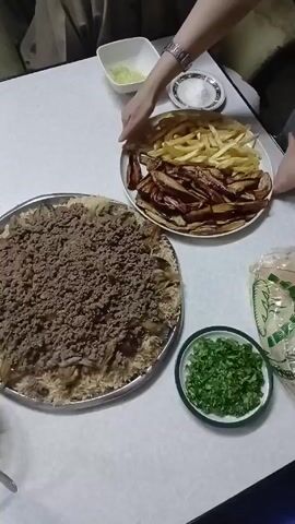 The video shows a table laden with food, set for a meal. There is a large plate of rice topped with ground meat, a plate with both regular and sweet potato fries, two bowls and a larger serving dish of chopped cucumber and tomato salad.  There are also small bowls with cut herbs and what appears to be a yogurt-based dip.  A stack of spoons is on the table. Two people are partially visible in the video, one reaching toward a bowl.