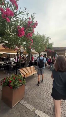 The video shows a paved area with a cobblestone surface and a wooden bench in the foreground. A large tree with pink flowers is on the left, with umbrellas and tables of an outdoor cafe visible behind it. In the background, a building with the text "STAZIONE LAGO" and "BIGLIETTERIA" is visible. People are walking around, some with backpacks, heading towards the building.

The subjects are a man and a woman walking away from the camera. The man is wearing a blue t-shirt, grey shorts, a baseball cap, and a black backpack. The woman is wearing a dark t-shirt, black shorts, and a black backpack. She has bright green and yellow sneakers. They appear to be walking towards the "STAZIONE LAGO" which likely indicates a lake station.

The scene appears to be a tourist area in Malcesine, Italy, given the text on the building. The weather seems to be overcast or slightly cloudy, but it's daytime. The overall mood appears casual and leisurely, as people are enjoying a day out in what looks like a charming European town. The presence of the cafe and the lake station suggests it is a public area with amenities for visitors. The text on the trash bin says "Comune di Malcesine".