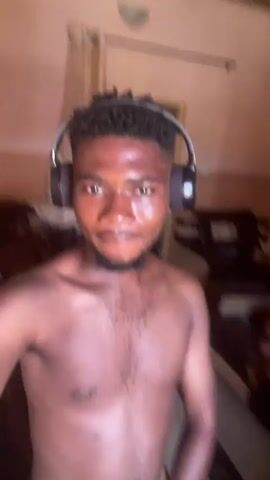 A shirtless dark-skinned man wearing headphones is the primary subject of this video. He has short, dark hair, a beard, and appears to be indoors. The background includes a bed with assorted items on it, a wall painted in a peach tone, a door, and a ceiling fan. The room seems to be sparsely furnished. The man initially looks directly at the camera, then shifts his gaze to the side before returning his attention to the camera.