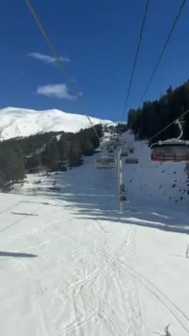 The video shows a view from a ski lift ascending a snowy slope. The sky is a clear, bright blue with a few scattered clouds. Below, the slope is covered in snow with visible ski tracks. A line of ski lift chairs, some occupied by people, moves upwards towards the top of the mountain. Trees dot the slope to the right, and a larger snow-covered mountain rises in the background. The ski lift cables extend upwards, converging towards the top of the frame.