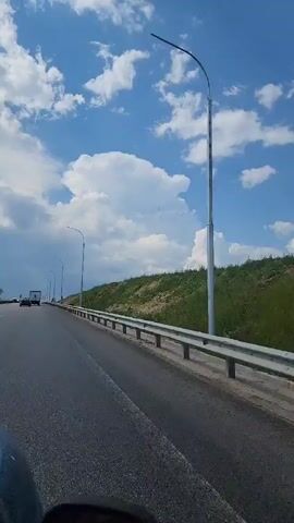 A first-person perspective video captures a view from a moving vehicle on a highway. The road stretches ahead, bordered by a metal guardrail and a grassy embankment. The sky is a vibrant blue, dotted with large, white, puffy clouds. A few cars and trucks are visible on the highway in the distance. Streetlights line the side of the road. The weather appears to be sunny and clear.