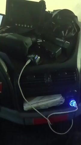 This is a first-person perspective video, likely from inside the cabin of a truck or commercial vehicle, as evidenced by the dashboard layout and a large display screen. The setting appears to be an interior space, possibly a truck cab, with various electronic devices and cables visible.

The subjects are primarily objects within the vehicle's cabin. These include a dashboard with illuminated displays, a steering wheel with a Mercedes-Benz logo, a touchscreen monitor, various buttons and switches, a USB charger with a blue light, and a bundle of papers. There are also cables, including a white USB cable, and what appears to be a bottle of water in the background.

The activity depicted is the general operation or presence within the vehicle's cabin. The illuminated displays and charging devices suggest the vehicle is powered on or in use. The video seems to be a panning shot, moving across the dashboard and interior of the vehicle, perhaps for documentation or personal record.

Notable details include the illuminated hazard light button (a pink triangle), various green indicator lights, and the digital displays on the dashboard showing information like speed (indicated by a needle gauge up to 120) and potentially other vehicle diagnostics. There's also text visible on the central display, though it's difficult to fully decipher. On one screen, there appears to be a circular gauge with a needle, possibly indicating some form of performance metric. The lighting suggests it could be daytime, or the interior lights are on.

No people are clearly visible in the video. There are no strong indicators of weather conditions, and the location cues are limited to the interior of the vehicle. No overt emotions are conveyed.

The visible text on the main display includes what appears to be Russian or a similar Cyrillic-based language, with phrases like "ВНИМАНИЕ СВЕТ" which translates to "ATTENTION LIGHT" and another line that seems to be an instruction or notification. The time stamp "00:00" is also displayed at the beginning of the video.