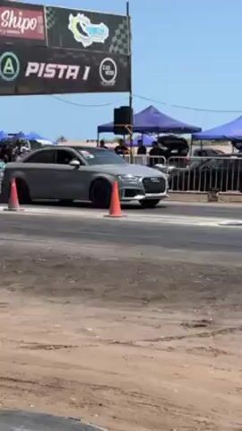 The video shows a gray Audi RS3 accelerating from the starting line of a drag race. The car is a four-door sedan with black wheels. The race takes place on a paved road with a dirt shoulder. There are various tents and vehicles visible in the background. An inflatable arch marks the starting line. Two men are standing near the starting line. There are cones and tires along the side of the track. The sky is blue and clear.