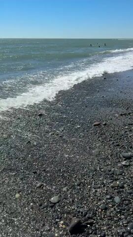 A low-angle shot of a pebble beach on a sunny day. The ocean is calm with gentle waves breaking on the shore. In the distance, several people are swimming in the water, and a few more are visible on the beach further down. The sky is clear blue.