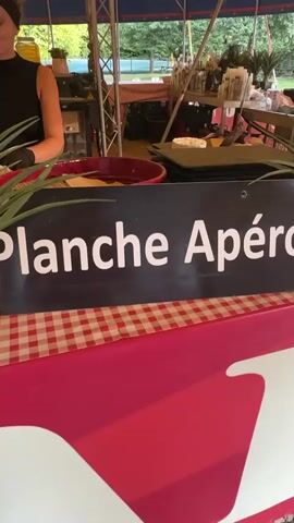 A person is standing behind a counter under a striped tent. The counter is covered with a red and white checkered cloth and has a sign that reads "lanche Apéro". There are various food items displayed on the counter, including cheese and sausage. In the background, there are more tables with food and drinks, as well as some potted plants. Another person is visible on the right side of the frame, interacting with the counter. The scene appears to be an outdoor market or food stall.