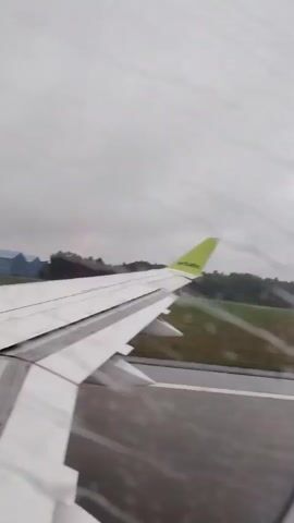 A static camera, looking out of an airplane window, captures the right wing of the plane as it moves along a tarmac, with green grass and trees in the distance under a cloudy sky.
