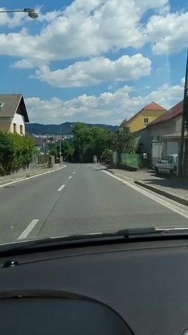 The video shows a first-person view from inside a car driving down a paved road. The road is lined with houses and trees on both sides, with a view of hills and a town in the distance. The sky is blue with fluffy white clouds, suggesting a pleasant, sunny day, likely in the late morning or early afternoon.

The houses appear to be residential, with some having colorful facades. There are fences and vegetation alongside the road, indicating a suburban or semi-rural setting. The road itself is marked with white lines, and there are streetlights visible.

The activity taking place is a car journey. The camera is positioned to capture the road ahead, the dashboard of the car, and the passing scenery. There are no people clearly visible in the foreground, but there might be figures in the distance.

No specific text is clearly legible in the video. The overall mood is calm and observational, typical of a casual drive. The location context provided suggests the drive is taking place in Písek, Czechia.