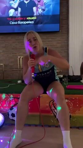 A young woman with blonde hair is singing into a red microphone. She is wearing a dark blue leotard with a light blue and yellow design on the front, and white socks. She is sitting on a multi-colored platform that has soccer ball designs on it. There are lights and what appears to be green turf behind her. A large screen is mounted on a brick wall behind her, displaying lyrics to a song and an image of four men. The lighting is dim, with colorful lights from the platform illuminating the woman.