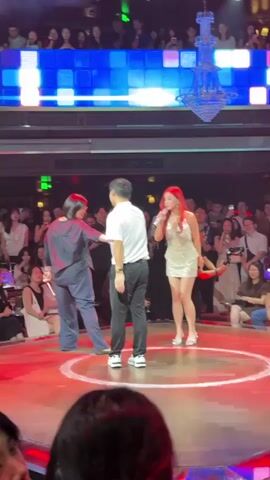 A woman with red hair in a sparkly dress is singing into a microphone on a circular stage. She is wearing silver high heels. A man in a white polo shirt and black pants stands facing her, with his back to the camera. To the left of the man is a person in a dark blue outfit, facing the woman and facing away from the camera. There is an audience seated around the stage, many of whom are holding up their phones, possibly recording. Above the audience is a wall with colorful flashing square lights and a large crystal chandelier. The stage floor is wooden and has a circular pattern. The overall setting appears to be a dimly lit nightclub or performance venue.