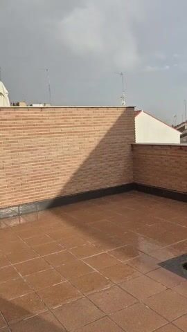 The video shows a close-up, eye-level, stationary shot of a wet, tiled rooftop with a brick wall on the left and a smaller brick wall on the right. There is a drain cover in the bottom right corner of the frame. The sky is overcast with clouds. There are houses and antennas visible in the background. There are small white objects falling from the sky, possibly rain or hail.