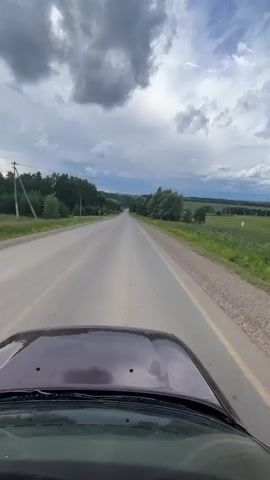 The video shows a first-person view from inside a car driving on a paved road. The car's hood is visible in the foreground. The road extends into the distance, flanked by grassy fields and trees. The sky is cloudy.