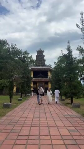 A group of tourists walks down a red brick pathway towards a yellow building with a multi-tiered pagoda on top. The pathway is lined with green grass and trees. The sky is cloudy.