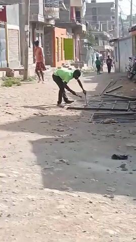 The video shows a man welding a metal structure on a dirt road in what appears to be a residential or commercial area. The man is wearing a green shirt and dark pants, and he is bending over the metal structure, using a welding tool. The metal structure appears to be a gate or a fence. There are other people walking in the background, and there are buildings on either side of the road. A motorcycle is parked on the right side of the road. The environment appears dusty and somewhat rundown.