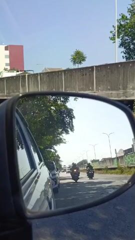 The video captures a view from inside a car, reflected in the side-view mirror. The mirror shows a road with moderate traffic, including several motorcycles and cars. Two motorcycles are prominently visible in the mirror, with riders wearing helmets. In the background, there is a concrete overpass or bridge structure, trees, and a building with a red facade. The sky is clear and blue. The text superimposed on the video is in Indonesian, and translates to "Study at a Superior Campus, a campus for champions with 14 scholarship programs." This suggests the context of promoting an educational institution.