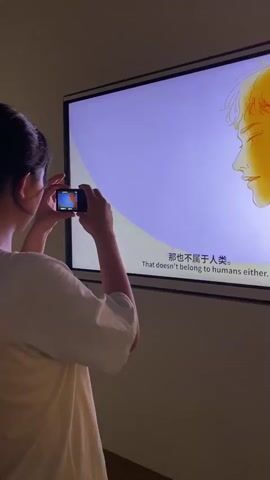A person wearing a white t-shirt films a large screen with a small digital camera. The screen displays animated graphics. In the first few seconds, the animation shows a profile of a person's face and a large purple circle with yellow accents. Then, the image transitions to a figure meditating in silhouette, surrounded by delicate lines and a blooming lotus flower that glows with bright yellow and orange light. The animation continues to focus on the glowing lotus flower. The person filming is standing with their back to the camera, holding the camera up to eye level and looking at the screen. The environment appears to be dimly lit, with the screen being the primary light source.