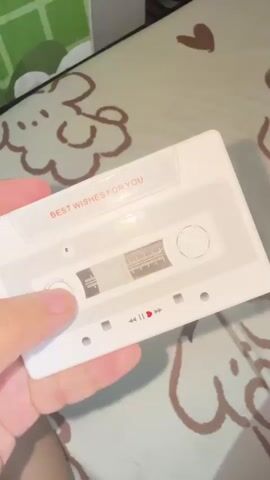 This media file shows a close-up shot of a white cassette tape with the text "BEST WISHES FOR YOU" printed in orange on its label. The cassette tape is held by a person's hand, with their thumb and index finger visible. The tape has a transparent window showing the magnetic tape inside, and there are play, pause, and fast forward icons printed below the window. The background appears to be a bed with a patterned sheet featuring cartoon-like cloud and heart designs. The lighting suggests an indoor setting, possibly during the daytime. The overall tone is casual and personal. There are no specific location cues that would pinpoint Manila, Philippines, other than the context provided.
