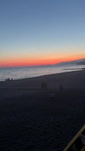 A video taken at sunset shows a tranquil beach scene. The sky displays a gradient of colors, from a deep blue at the top to a vibrant orange and pink band just above the horizon, fading into the distant mountains. The sea is calm, with gentle waves lapping at the shore. The beach is dark and pebbly. Several people are visible in the distance, appearing as small silhouettes. Closer to the foreground, two children are playing on the sand. The camera appears to be held by someone in the foreground, partially obscuring the lower right corner with a yellow and black object. The overall atmosphere is peaceful and serene.