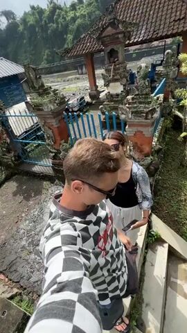 In this video, a man wearing sunglasses and a black and white checkered long-sleeved shirt is captured from a high-angle perspective as he descends a set of concrete stairs. He is accompanied by a woman, partially visible, wearing a white skirt and a patterned top. Behind them, a traditional Balinese temple structure with a tiered roof is visible. The temple is adorned with intricate carvings and features blue fencing. Lush green vegetation surrounds the temple and cascades down a slope towards a river. The river itself appears somewhat turbulent, with a concrete dam or weir visible in the background, creating a whitewater effect. The overall setting suggests a warm, possibly humid climate, typical of a tropical region like Bali. The man's gaze is directed downwards, and the woman appears to be looking at something to her right. The scene unfolds during daylight, with dappled sunlight suggesting it might be late morning or early afternoon. The atmosphere appears relaxed, as if the individuals are enjoying a scenic exploration. No visible text is present in the media.