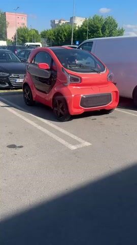 The video shows a small red car parked in a parking lot on a sunny day. The car has a unique design, with its door open. Other cars are parked nearby, including a black Audi and a white van. The parking lot is paved with asphalt, and there are some buildings and trees in the background.