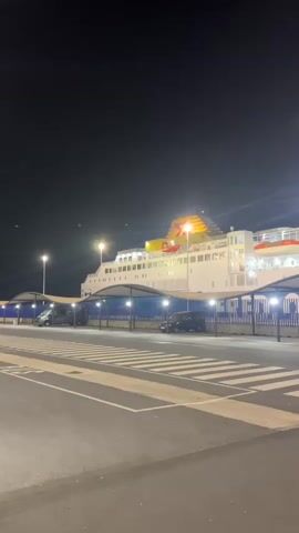 This is a vertical, nighttime video of a large white ferry boat docked at a port. The ferry has multiple decks and is illuminated by various lights, including yellow lights on its upper structure and white lights along its sides. Two lifeboats, painted orange and white, are visible on one of the upper decks. The ferry appears to have the name "FRED OLSEN" written in blue letters across its side, partially obscured by a covered waiting area. Beneath the waiting area, which has a blue and white striped fence, are several parked vehicles, including a dark SUV and a lighter-colored car. The foreground of the video shows a paved area with white pedestrian crossing lines, indicating a road or walkway leading to the port. The sky is dark, and there are several lampposts providing additional illumination to the scene.