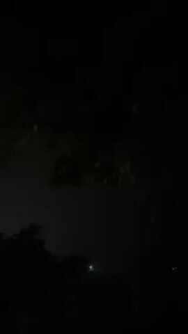 The video shows a nighttime scene with trees in silhouette against a dark sky.  A flash of light illuminates the scene, changing the sky to a purple hue.  Another flash occurs, and then the scene returns to darkness.  A faint light can be seen in the distance.  The sound of thunder can be heard.