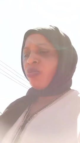 A woman wearing a black hijab and a white tunic with silver trim is looking at the camera. She is outdoors in a sunny area with buildings and power lines in the background.