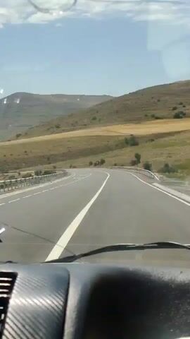 The video shows a view from inside a vehicle, looking out the windshield at a two-lane asphalt road curving through a dry, hilly landscape under a clear blue sky. The road has white lane markings and a metal guardrail on the left side. The hills are covered in dry grass and sparse vegetation, with some visible terracing on one of the larger hills in the background. The dashboard of the vehicle is partially visible in the foreground, with a textured surface.