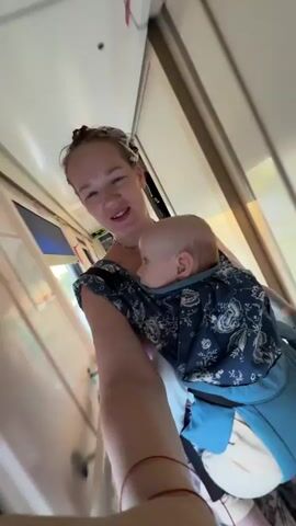 In this video, a woman is seen holding a baby in a blue and white floral carrier. The woman has her hair pulled up in a bun and is wearing a necklace. She smiles at the camera as she walks, with the baby facing outwards. The scene appears to be inside a train, with the interior of the train carriage visible. The woman moves down a narrow passageway, and the camera briefly captures a view out of a window.

The view from the window shows train tracks and a station platform. In the background, there are trees, a water tower, and a building. The lighting suggests it is daytime, possibly late afternoon, with bright sunlight. The overall mood is casual and perhaps indicative of travel. There is no visible text in the video. The location context provided is Uzunovo, Russia.