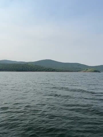 The video shows a wide view of a large body of water with a distant shoreline. The water is dark and appears to be slightly choppy. The shoreline is lined with a dense green forest that covers rolling hills. The sky is a pale blue with some hazy clouds. The camera seems to be moving slightly, giving the impression of being on a boat or a moving platform. There are no people or man-made objects visible in the frame. The overall impression is one of natural, undeveloped landscape.