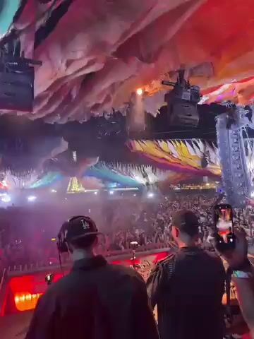 This video captures a high-energy moment at a large music festival or concert, viewed from behind the DJ booth. Two male DJs are the main subjects, standing with their backs to the camera, actively performing for a massive crowd. One DJ wears a black cap with "LA" visible on it, and he is seen raising his arm to the crowd, indicating engagement and excitement.

The scene is characterized by a vibrant and dynamic light show, featuring colorful lasers, spotlights, and smoke/haze filling the air above the audience. The ceiling of the venue is an elaborate, organic, and perhaps cave-like structure, illuminated with changing lights, predominantly red, yellow, and blue, creating an immersive atmosphere. Below the DJs, the control panel is brightly lit with red, indicating active equipment.

The crowd below is a sea of people, tightly packed and extending far back into the venue, all facing the stage and bathed in the shifting stage lights. The overall ambiance is one of intense energy and excitement, typical of a major electronic music event. Given the location of Indio, United States, this is likely at the Coachella Valley Music and Arts Festival. The time of day is clearly night or a dark indoor setting, optimized for the light show. A brief flash of text "end" appears on screen around 0:05.
