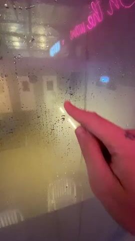 A close-up shot shows a hand with tattoos on a knuckle and fingers drawing on a steamy window. The person's index finger is visible, smudging condensation on the glass. In the background, neon lights glow, with the words "The Maze" in pink neon visible at the top of the frame. The drawing starts with two vertical lines for eyes and a horizontal line for a mouth, forming a basic smiley face. The finger then smudges the mouth, turning it into a wavy line, creating a more surprised or concerned expression. The overall scene is dimly lit, with the yellow and pink neon lights reflecting on the wet glass, creating an atmospheric and somewhat moody setting.