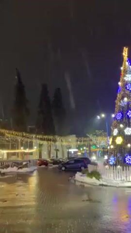 The video shows a decorated Christmas tree at night. The tree is lit with blue and yellow lights, and adorned with snowflake ornaments. The base of the tree is surrounded by a white picket fence. The ground is wet and there is snow falling. In the background, there is a building decorated with lights, and several cars are parked nearby.