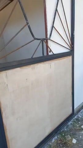 The video shows a person's hands opening a metal gate. The gate has a decorative wrought iron design at the top and a plain, light-colored panel in the lower section. The gate is attached to a white wall. The environment appears to be outdoors, with gravel and some debris on the ground. A wooden workbench or shelf unit is visible in the background. The lighting suggests it is daytime.
